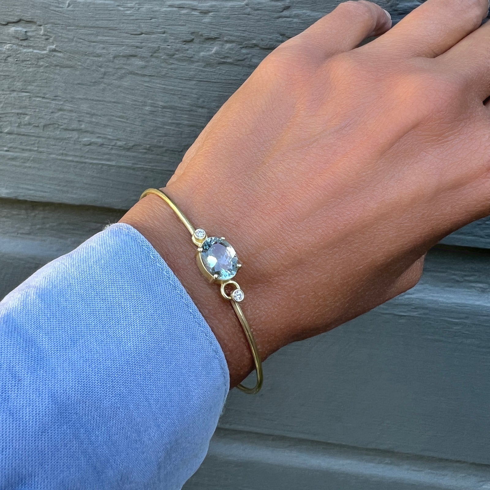 A gold bangle bracelet with a light blue oval aquamarine and a small diamond accent stone, shown worn on a person's wrist against a gray wooden background.