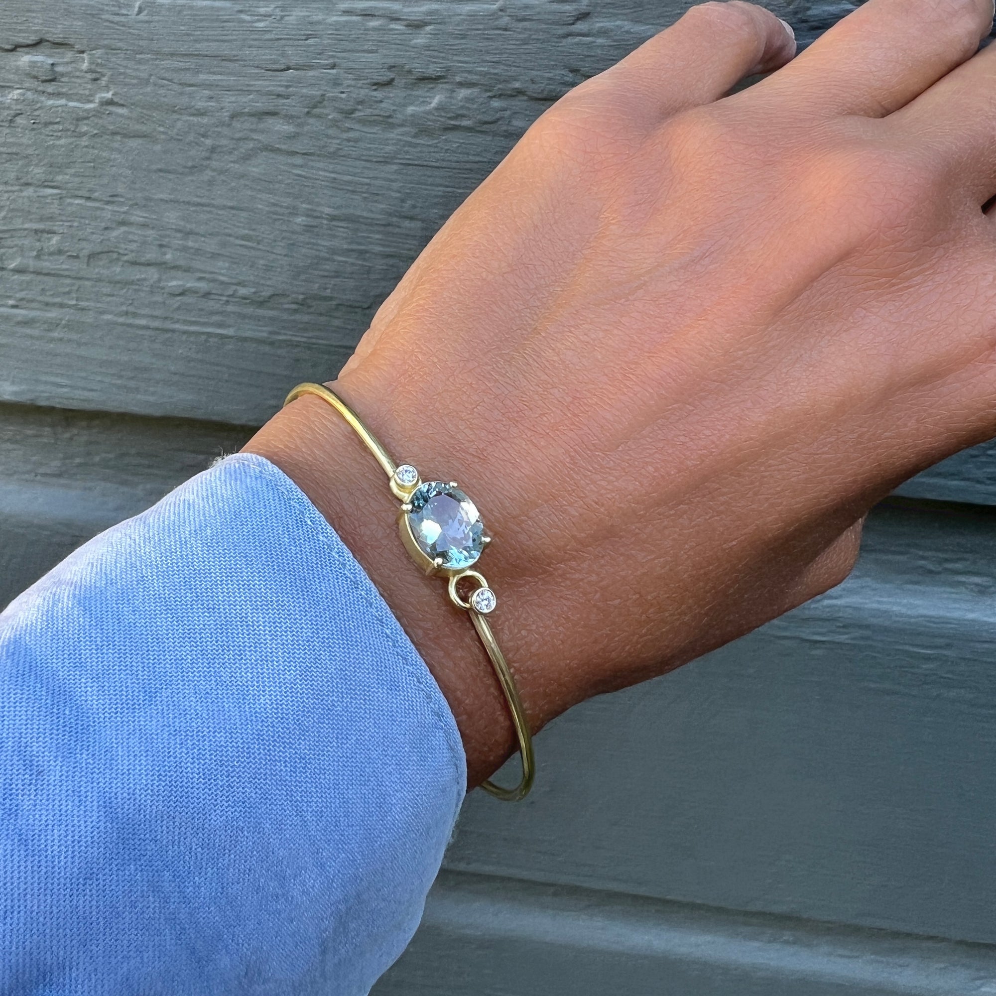 A gold bangle bracelet with a light blue oval aquamarine and a small diamond accent stone, shown worn on a person's wrist against a gray wooden background.