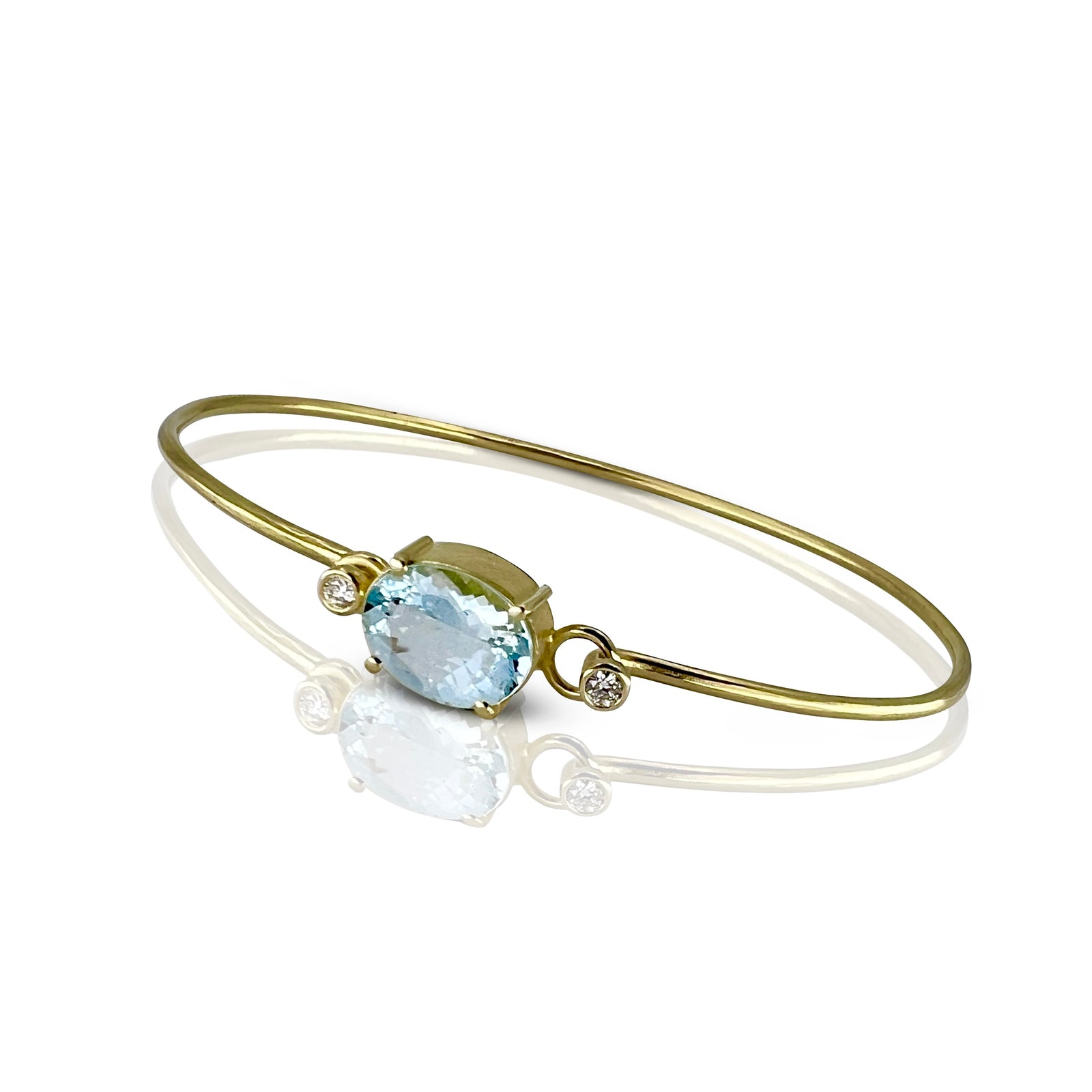 A gold bangle bracelet with a light blue oval aquamarine and a small diamond accent stone, shown worn on a person's wrist against a gray wooden background.